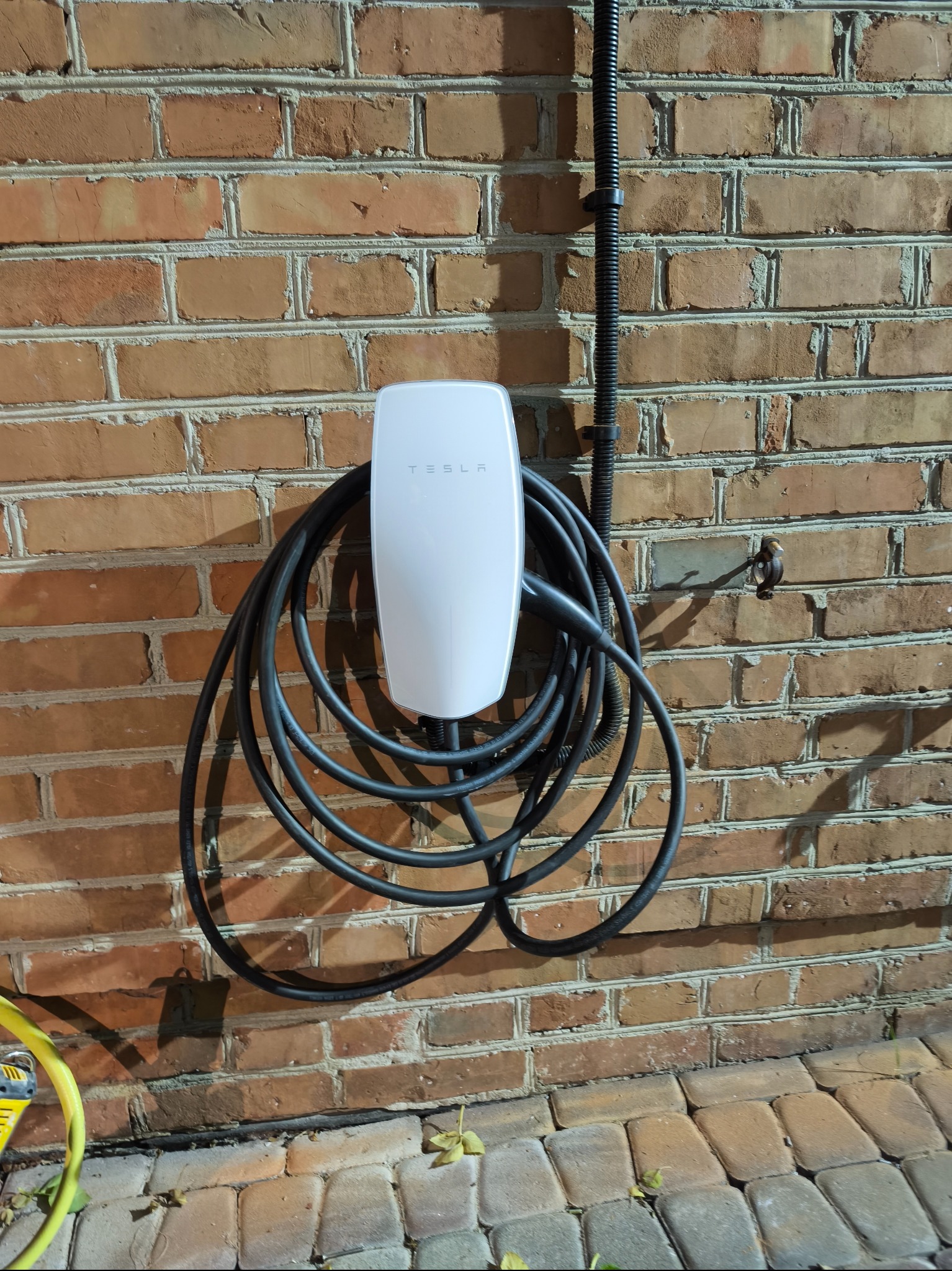 Biała ładowarka Tesla Wall Connector zamontowana na ceglanej ścianie zewnętrznej, z uporządkowanym kablem i widoczną rurą ochronną. Oświetlenie wieczorne.