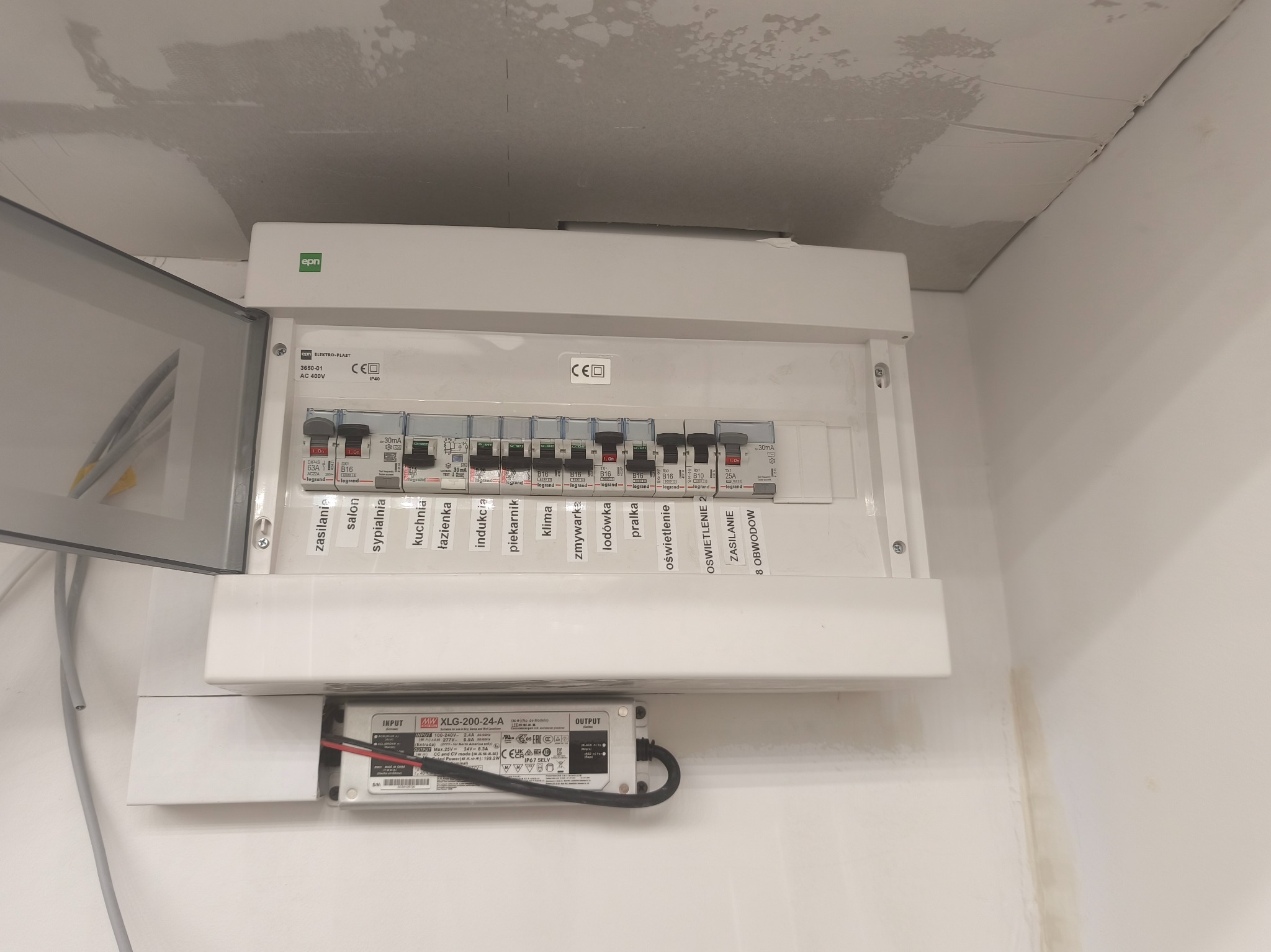 Otwarta skrzynka elektryczna z bezpiecznikami Legrand, opisana dla różnych obwodów (salon, kuchnia, oświetlenie), zasilacz LED XLG-200-24-A poniżej. Widoczne elementy instalacji.