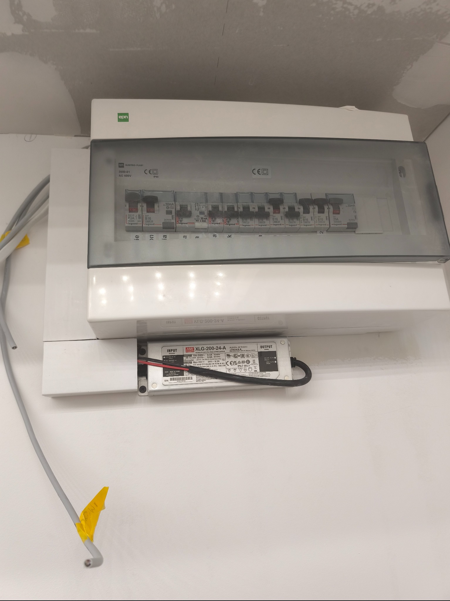 Biała skrzynka elektryczna z bezpiecznikami Legrand i zasilaczem Mean Well XLG-200-24-A, zamontowana na ścianie. Widoczne przewody z żółtą taśmą.