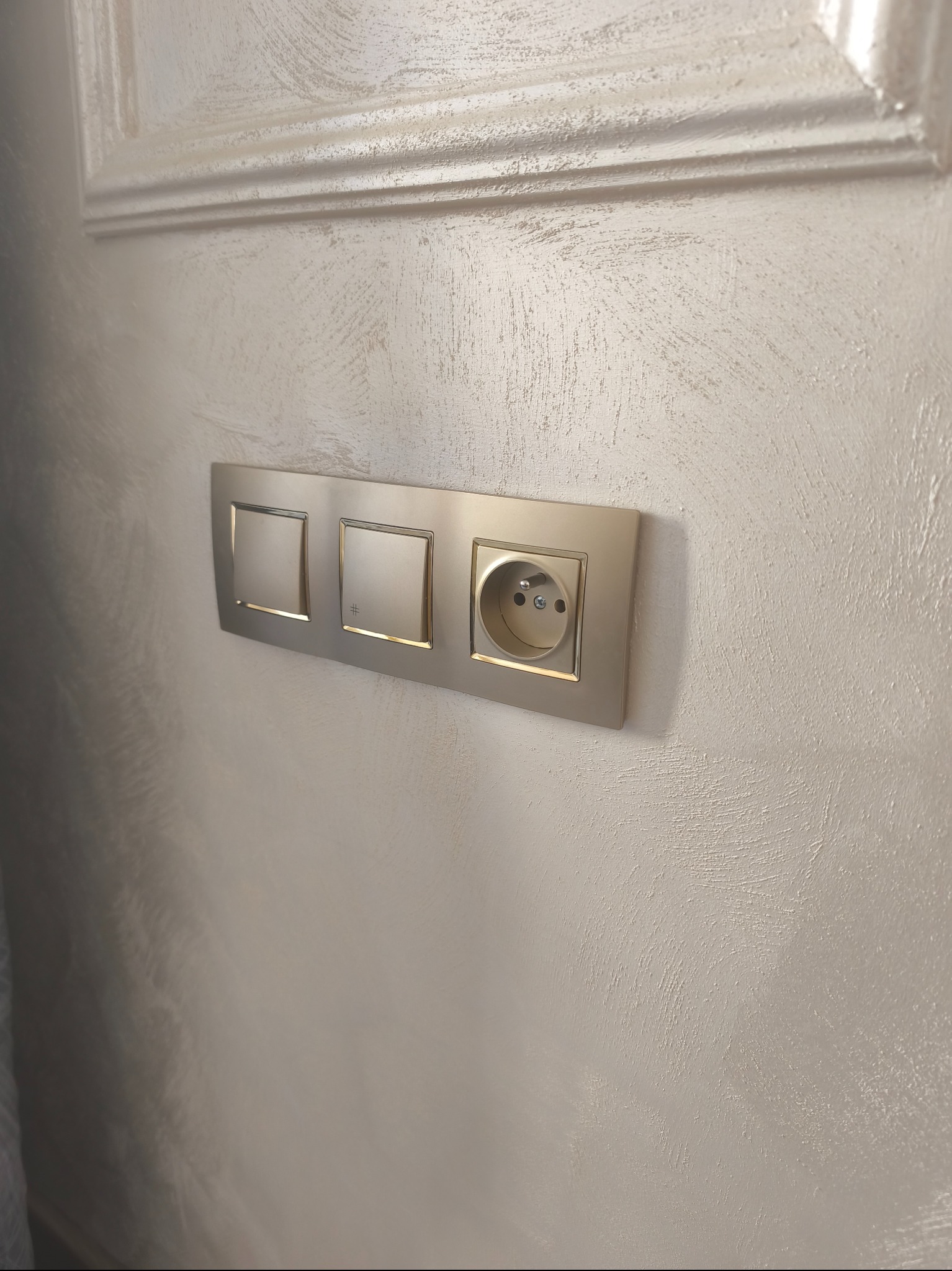 Zestaw gniazdka i włączników w ramce, zamontowany na ścianie o dekoracyjnej fakturze, pod ozdobnym elementem architektonicznym. Ujęcie z bliska, neutralna kolorystyka.