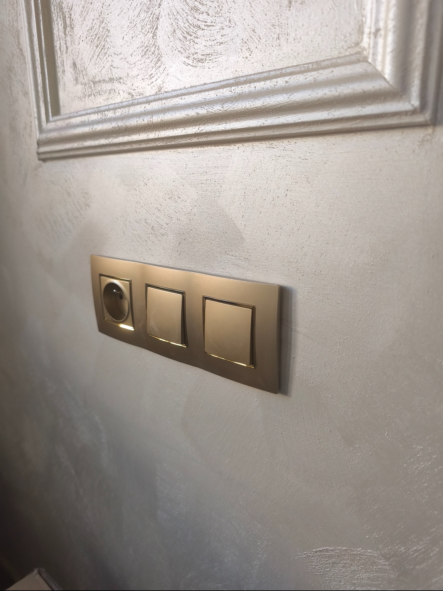 Złota listwa z gniazdkiem i dwoma włącznikami światła na ścianie z dekoracyjnym tynkiem, zamontowana pod ozdobną listwą przysufitową.