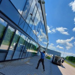 Clean Glass Professional - Mężczyzna myje szklaną fasadę budynku za pomocą lancy teleskopowej. Na chodniku leży wąż, a w tle widać zaparkowany samochód dostawczy i błękitne niebo.