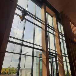 Clean Glass Professional - Pracownik alpinistyczny myje wysokie okna biurowca z zewnątrz, widok z wnętrza budynku na pochmurne niebo i krajobraz.