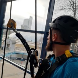 Clean Glass Professional - Pracownik w kasku i uprzęży, z narzędziami alpinistycznymi, spogląda przez okno na panoramę miasta. Widoczny fragment wejścia do metra.