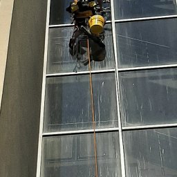 Clean Glass Professional - Pracownik na linie, w uprzęży, czyści okna wieżowca. Widoczne zabrudzenia na szybach i sprzęt do mycia. Ujęcie z dołu, w słoneczny dzień.