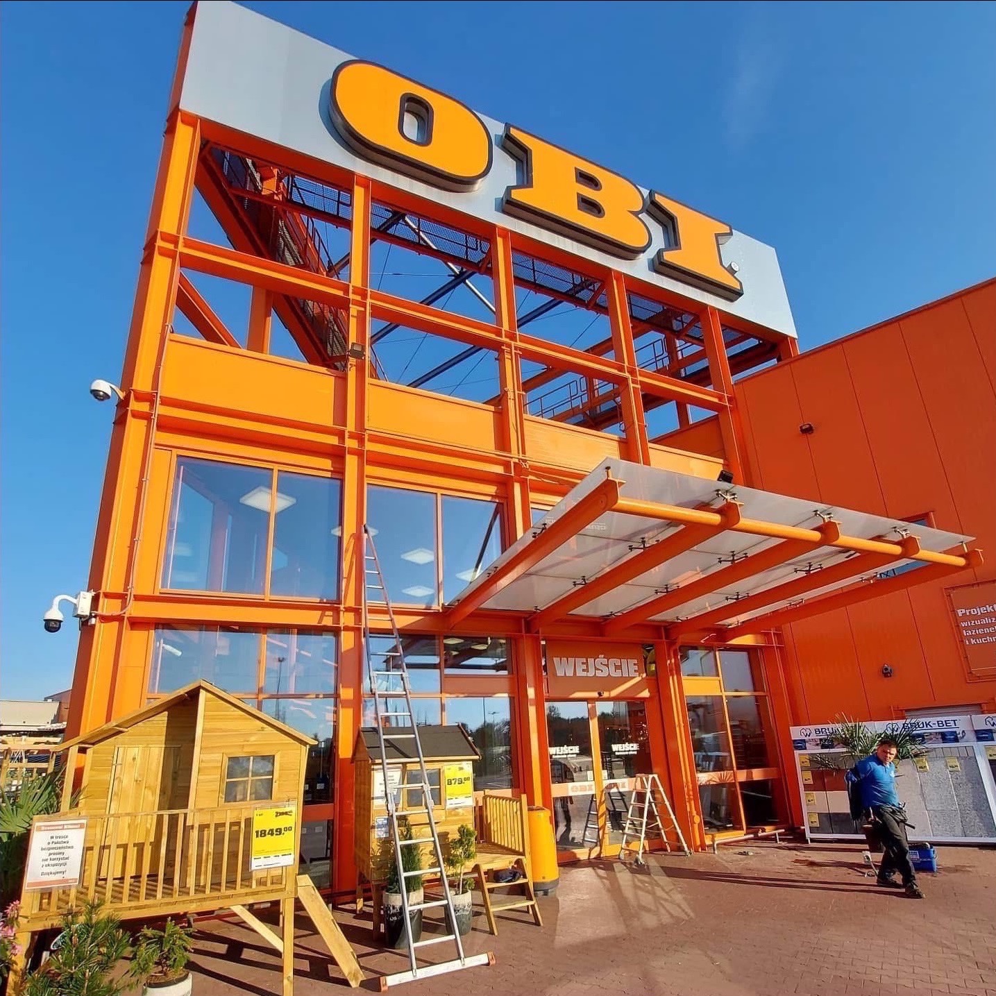 Pomarańczowy budynek OBI z drabiną opartą o okna, sugerujący prace na wysokości. Widoczny pracownik i ekspozycja domków dla dzieci.