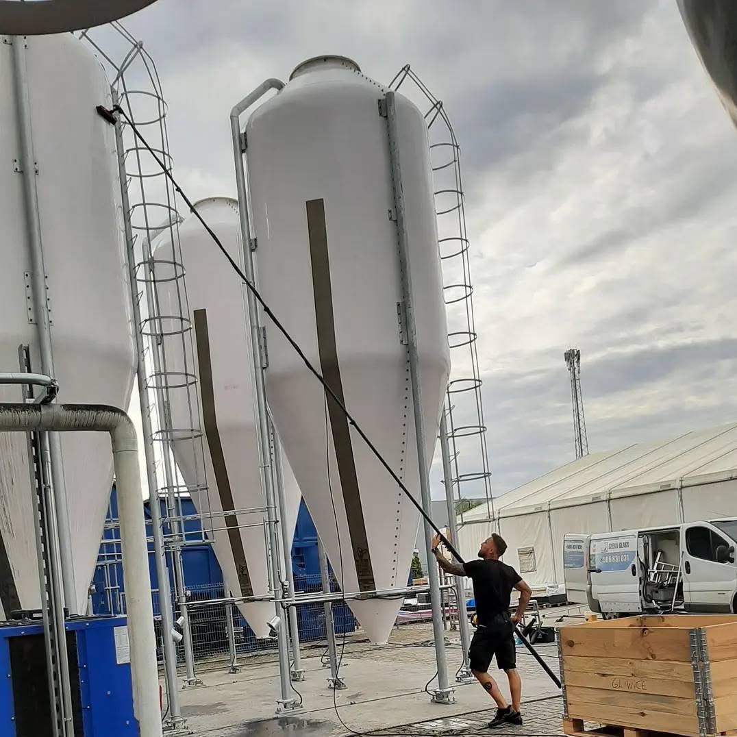 Mężczyzna myje wysokie, białe silosy z drabinkami za pomocą długiej lancy. Widoczne wozy serwisowe i skrzynia z napisem Gliwice. Pochmurne niebo w tle.
