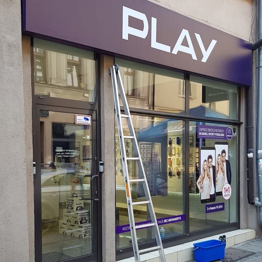 Drabina oparta o witrynę salonu Play przed myciem. Niebieskie wiadro i ściągaczka u dołu. Widoczne odbicie ulicy w szybach.