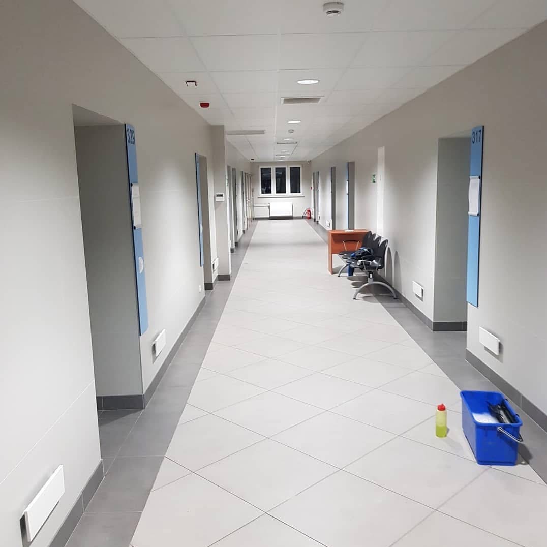 Długi, jasny korytarz z białymi ścianami i szarą podłogą. W oddali okno, a po prawej stronie widoczny niebieski mop i butelka środka czyszczącego.