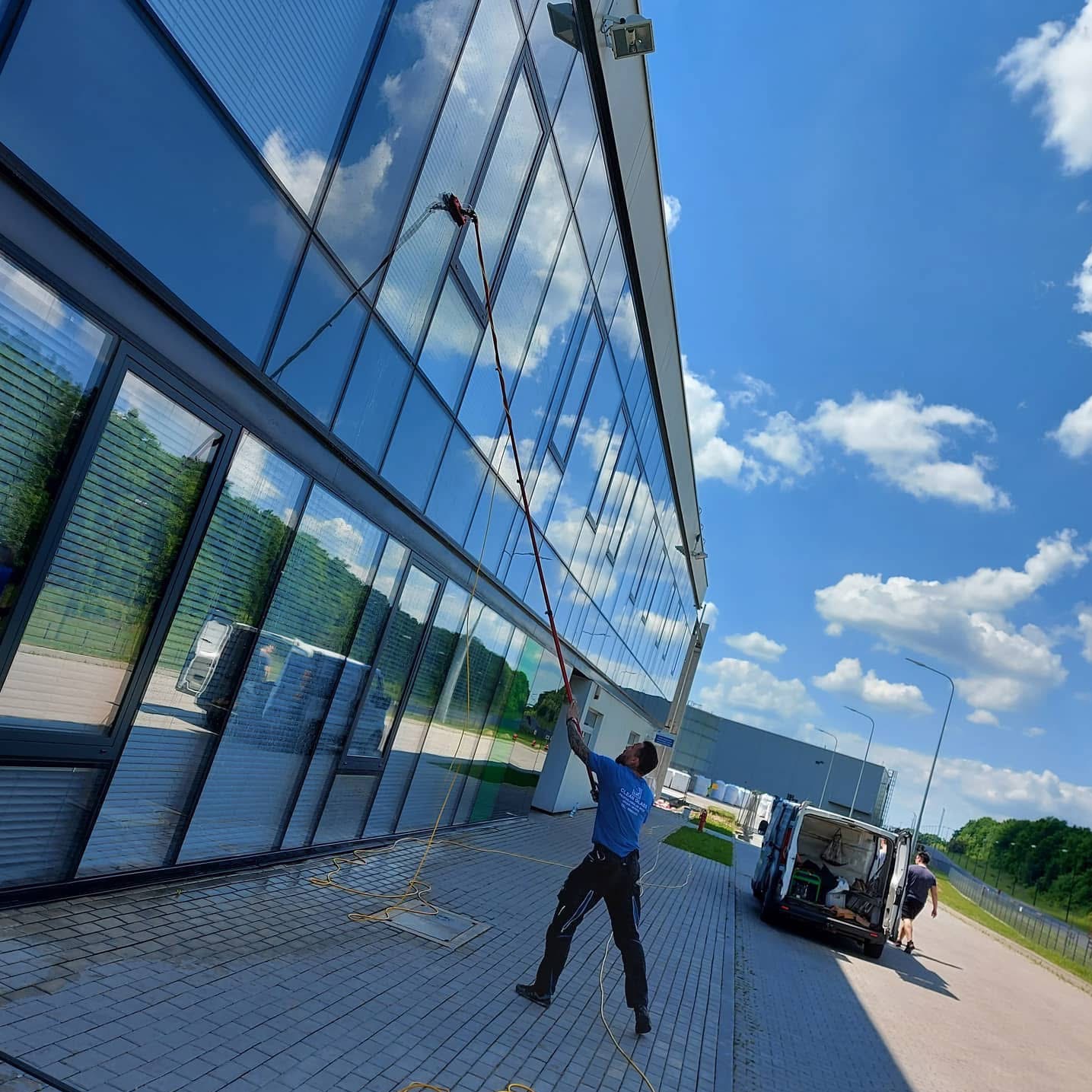 Mężczyzna myje szklaną fasadę budynku za pomocą lancy teleskopowej. Na chodniku leży wąż, a w tle widać zaparkowany samochód dostawczy i błękitne niebo.