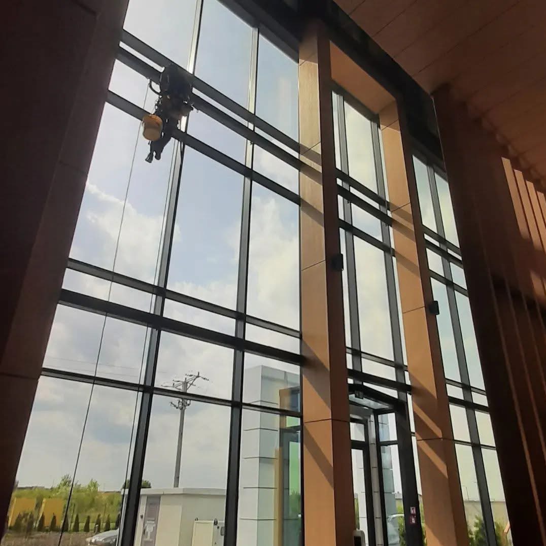 Pracownik alpinistyczny myje wysokie okna biurowca z zewnątrz, widok z wnętrza budynku na pochmurne niebo i krajobraz.