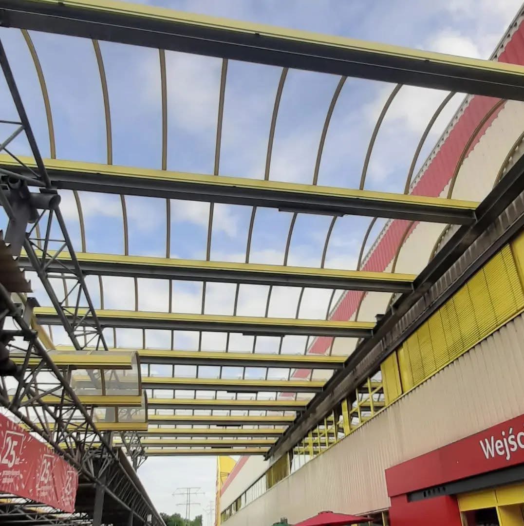 Zadaszenie z żółtymi elementami konstrukcyjnymi i przezroczystym wypełnieniem, widok z dołu, z fragmentem budynku w tle i elementami konstrukcji stalowej po lewej stronie.