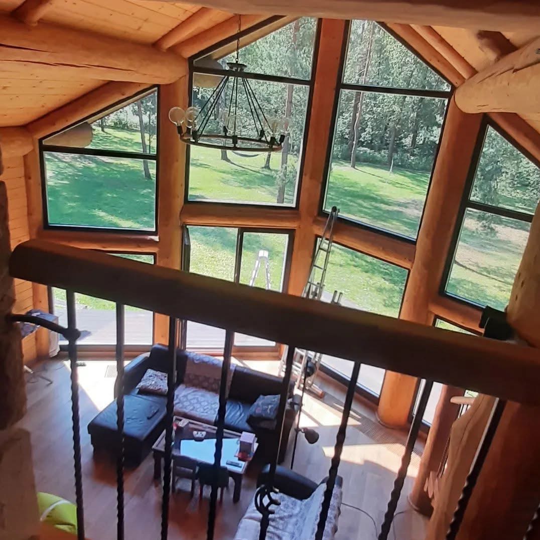 Wnętrze drewnianego domu z wysokimi oknami, przez które widać zieleń. Metalowa drabina oparta o okno, sofa i stolik w salonie, widok z góry zza balustrady schodów.