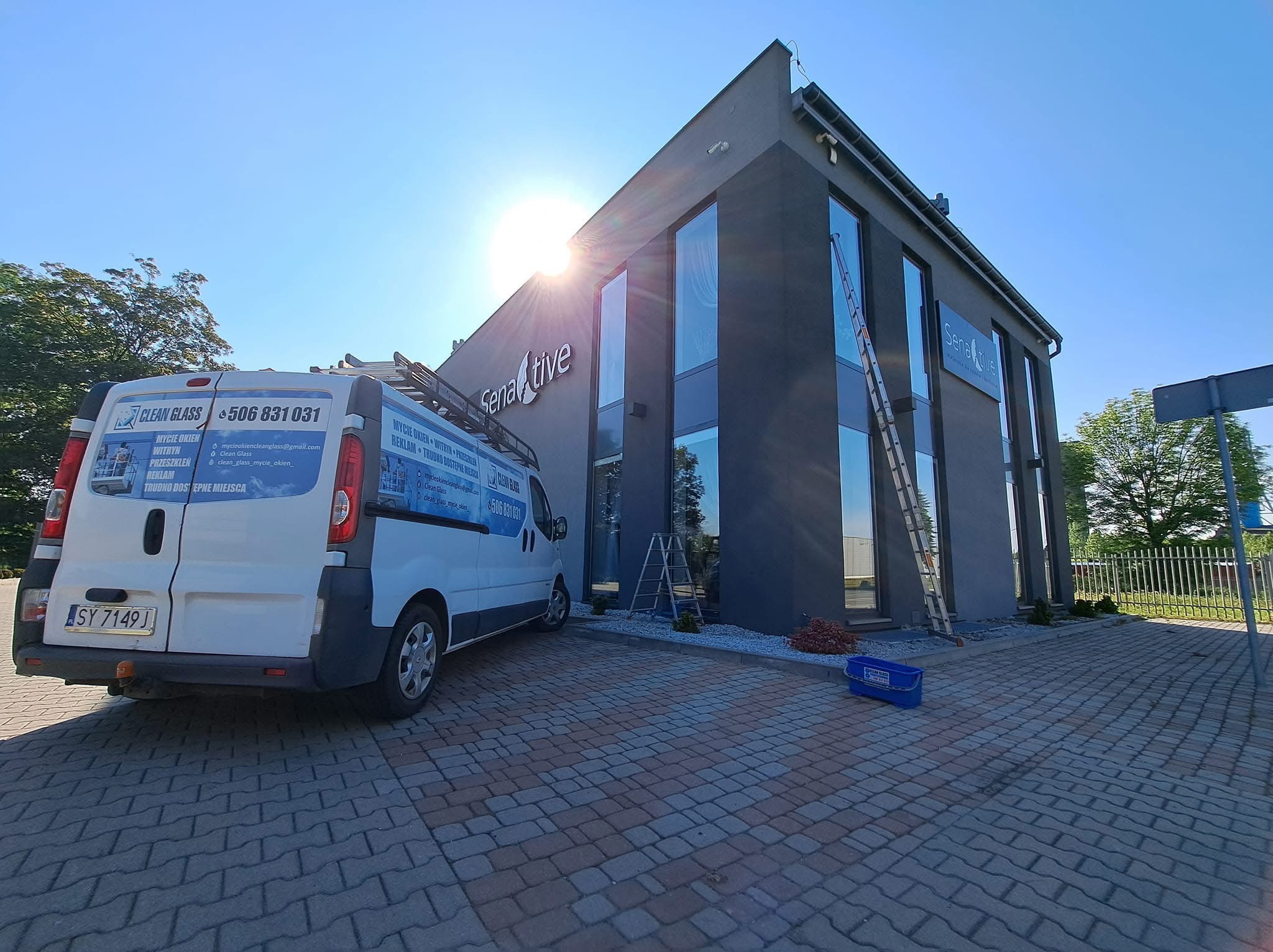 Mycie okien biurowca: budynek z dużymi oknami, drabina oparta o ścianę, bus firmy Clean Glass z numerem telefonu i informacjami o usługach, słońce w tle.