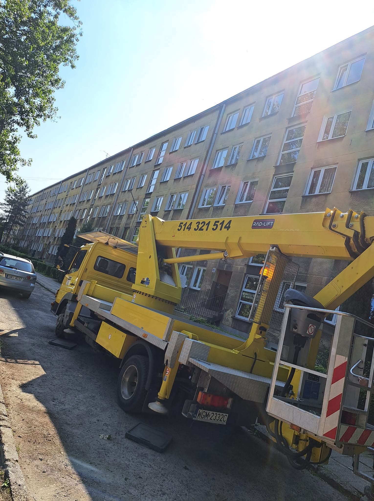 Żółty podnośnik koszowy RAD-LIFT na tle bloku mieszkalnego z licznymi oknami, zaparkowany na ulicy. Na wysięgniku widoczny numer telefonu. Słoneczny dzień.