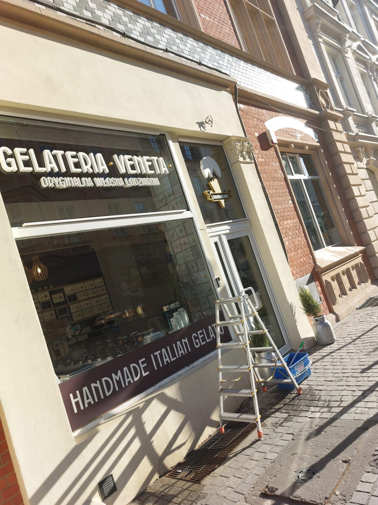 Aluminiowa drabina oparta o witrynę lodziarni 'Gelateria Veneta' z napisem 'Handmade Italian Gelato'. Widoczne wiadro i akcesoria do mycia okien. Słoneczny dzień.
