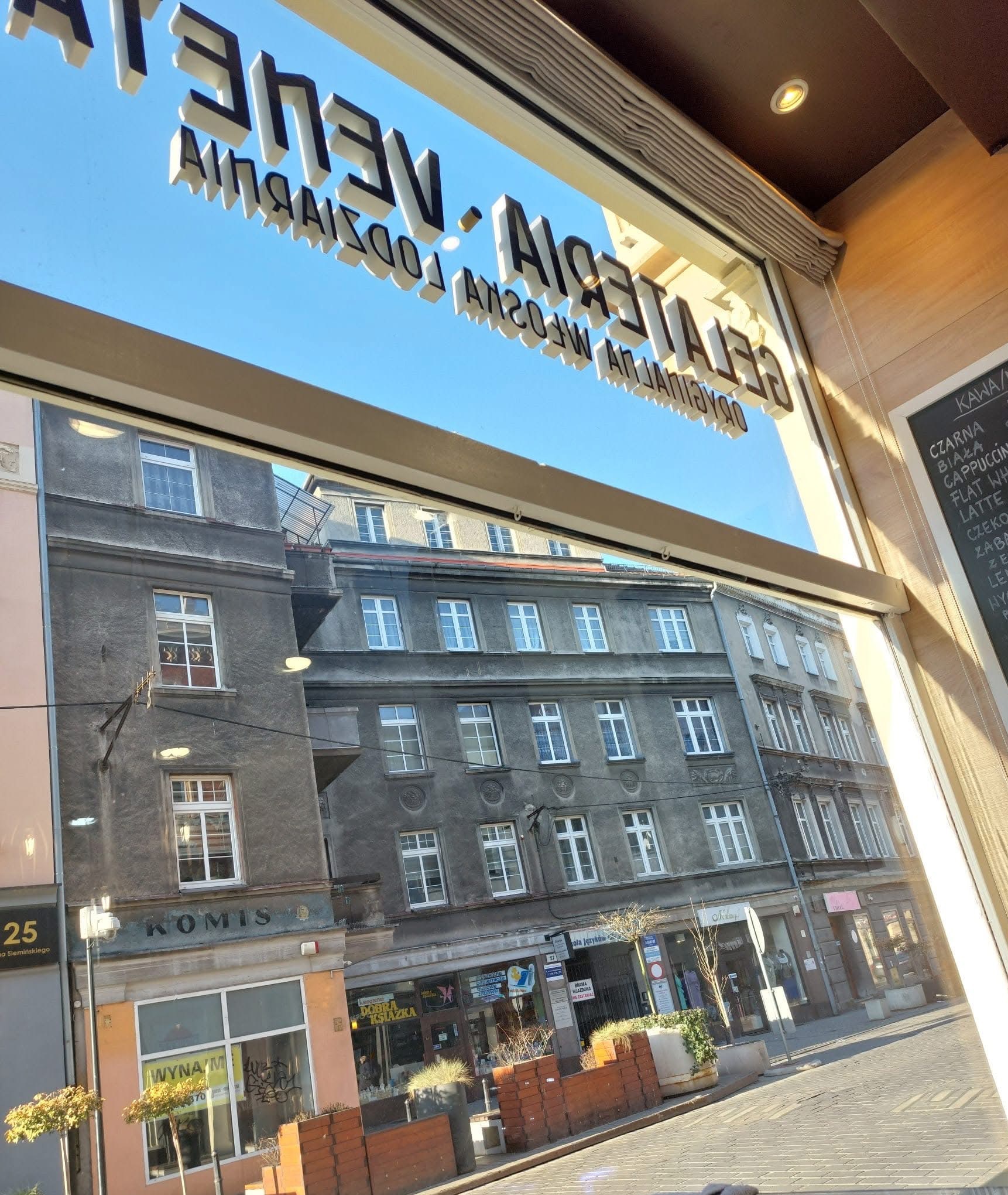 Widok z kawiarni na ulicę miasta przez okno z napisem 'Gelateria' i menu kawowe na tablicy. Słoneczny dzień, budynki w tle.