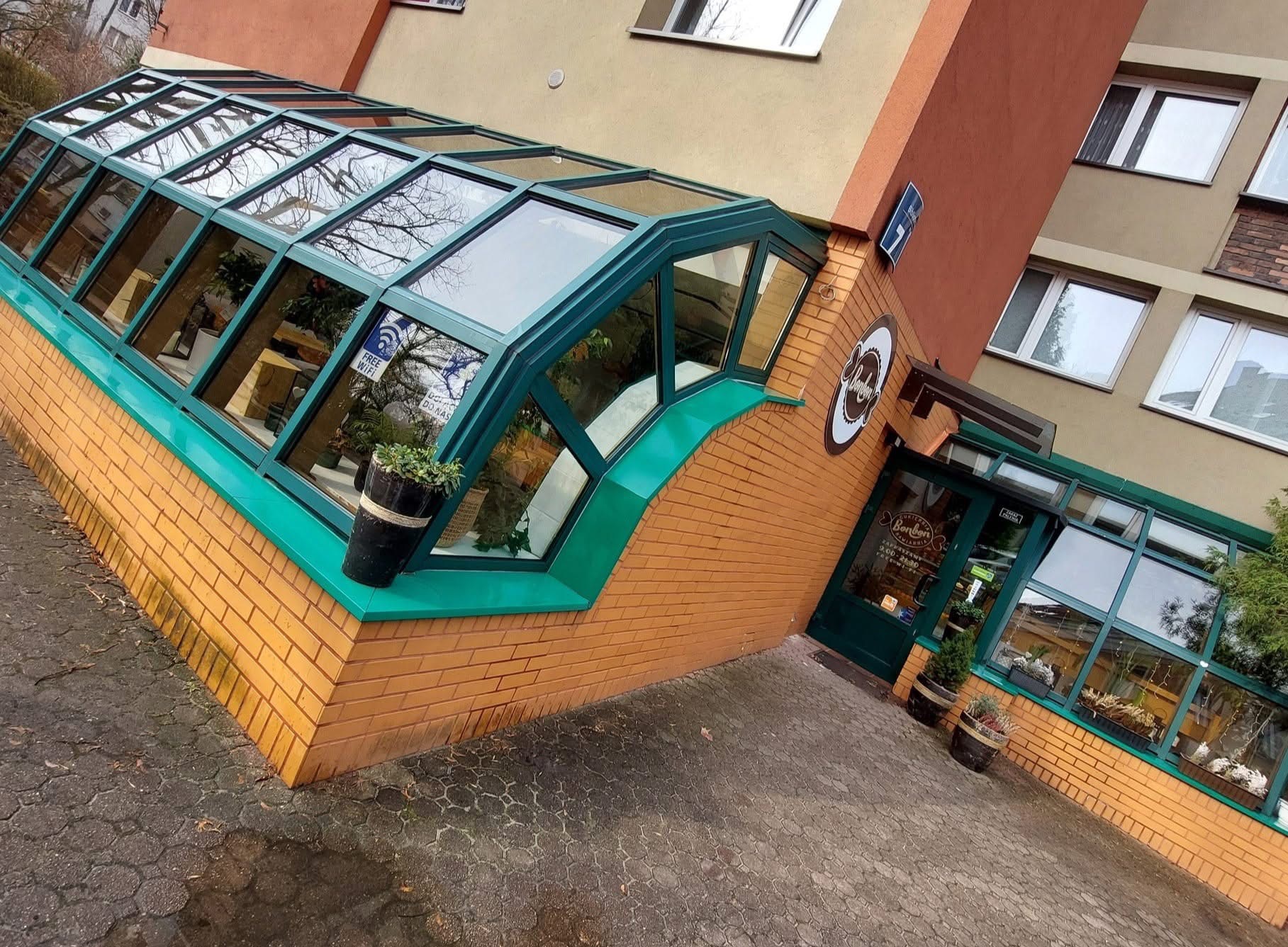 Fasada budynku z przeszklonym wejściem i zielonymi elementami, donice z roślinami. Widoczne okna budynku i fragment chodnika z kostki brukowej. Ujęcie pod kątem.