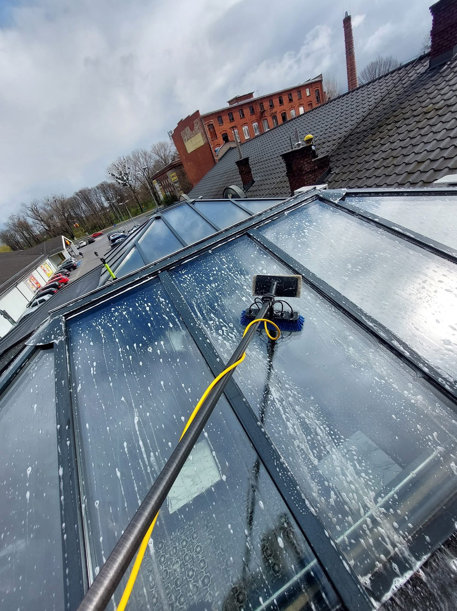 Mycie przeszklonego dachu narzędziem teleskopowym z żółtym przewodem. W tle budynek z czerwonej cegły i szare niebo. Widoczne pęcherzyki piany na szybach.