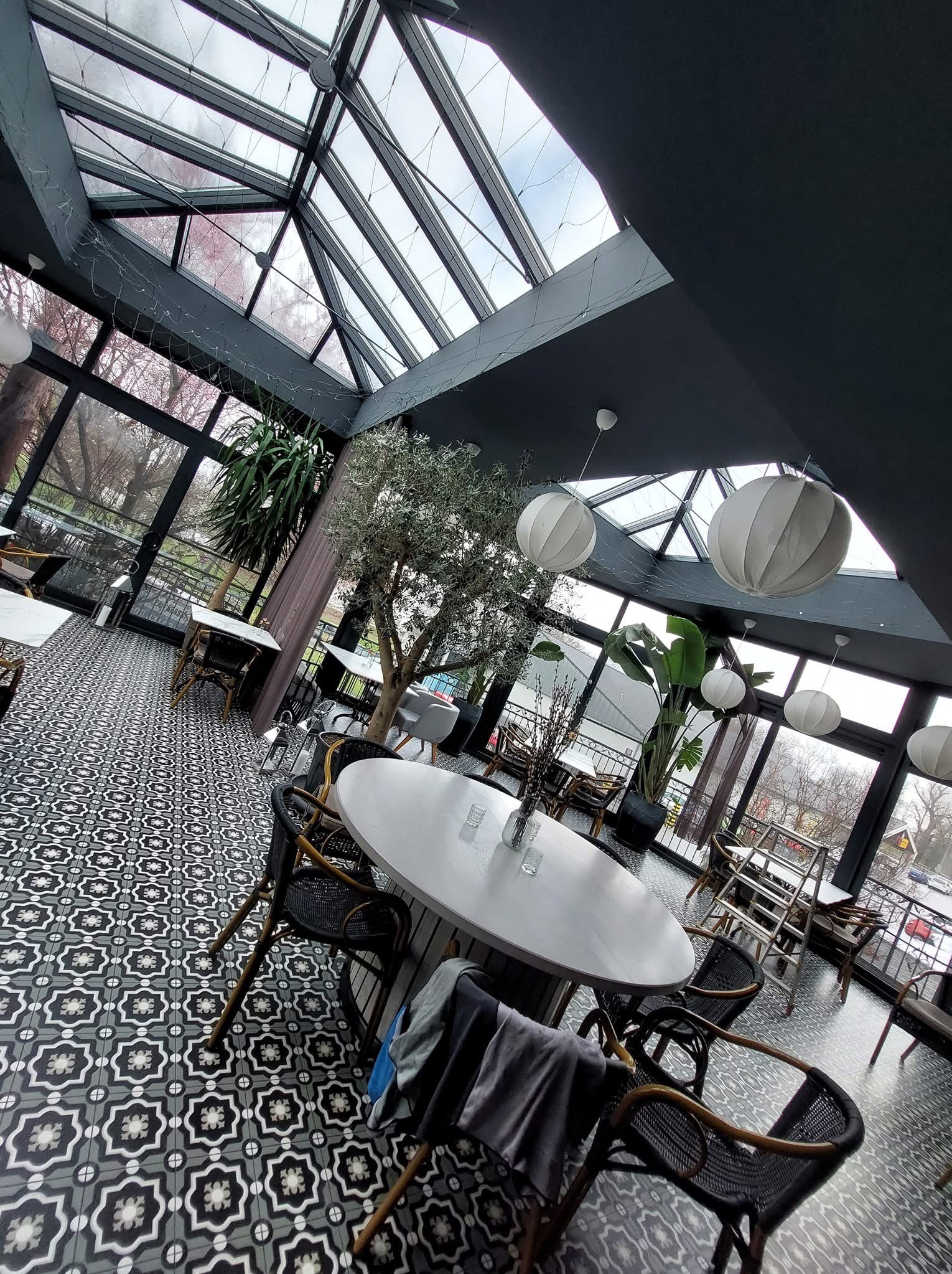Wnętrze oranżerii w Bytomiu z przeszklonym dachem, roślinami doniczkowymi i stolikami z czarnymi krzesłami. Dekoracyjna podłoga w geometryczne wzory.