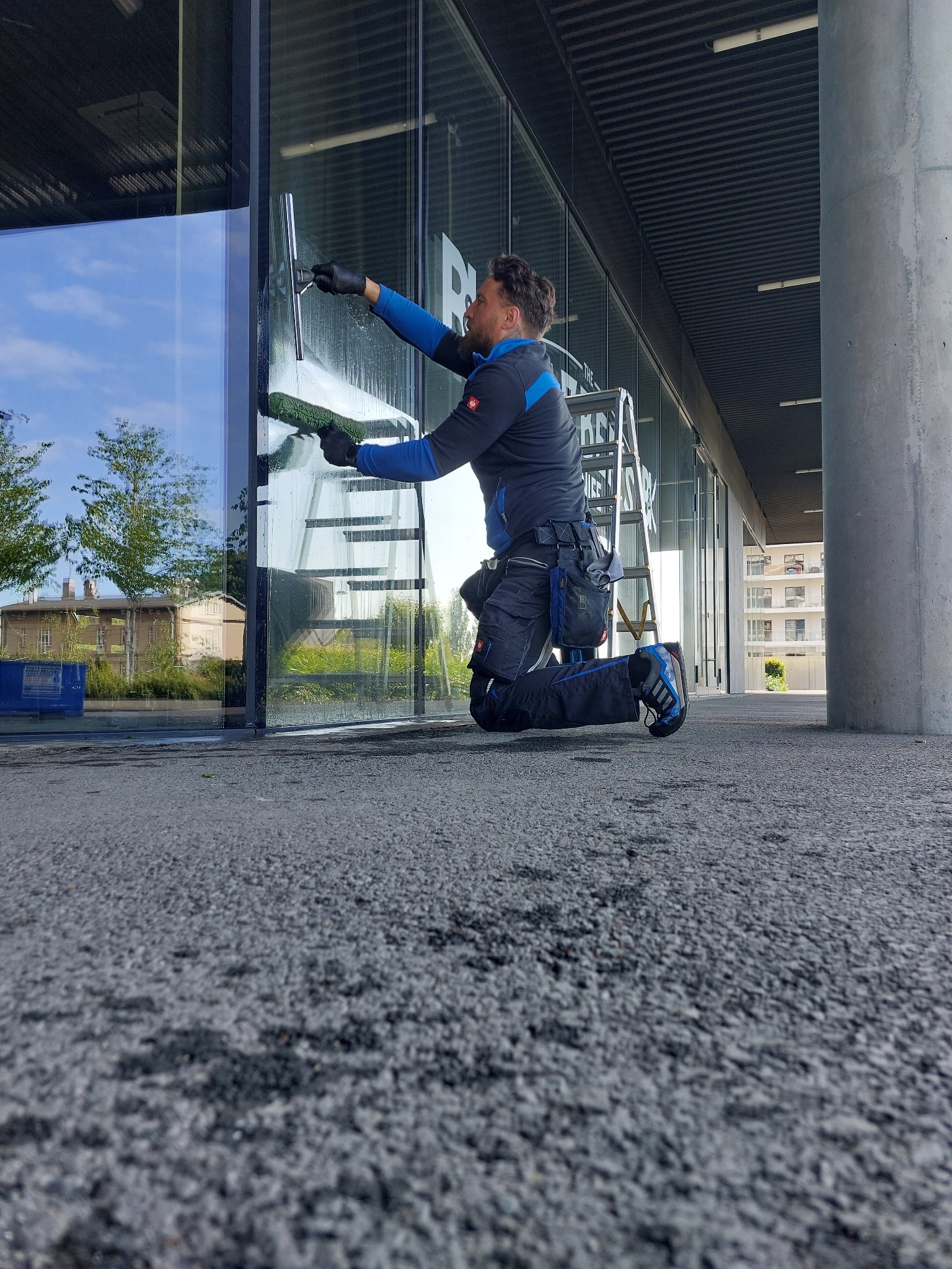 Pracownik w czarnych rękawicach czyści szklaną fasadę budynku za pomocą ściągaczki i myjki. Widoczna drabina, betonowy filar i budynki w tle.