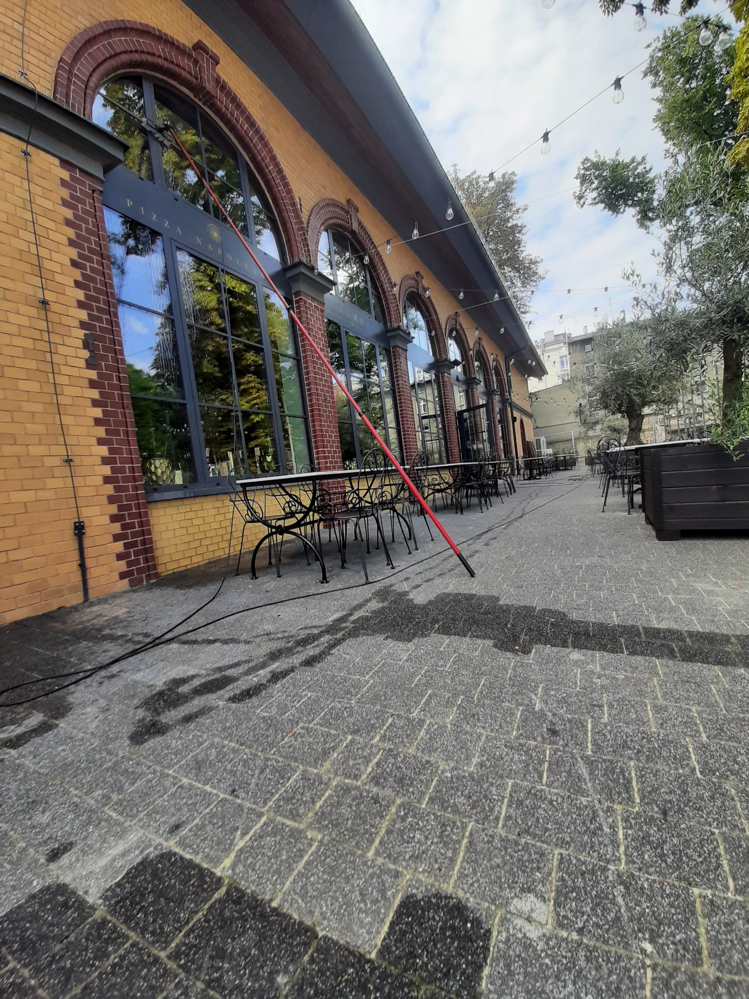 Mycie okien na zewnątrz budynku z cegły z użyciem lancy teleskopowej. Widoczne mokre ślady na kostce brukowej i odbicie drzew w oknach.