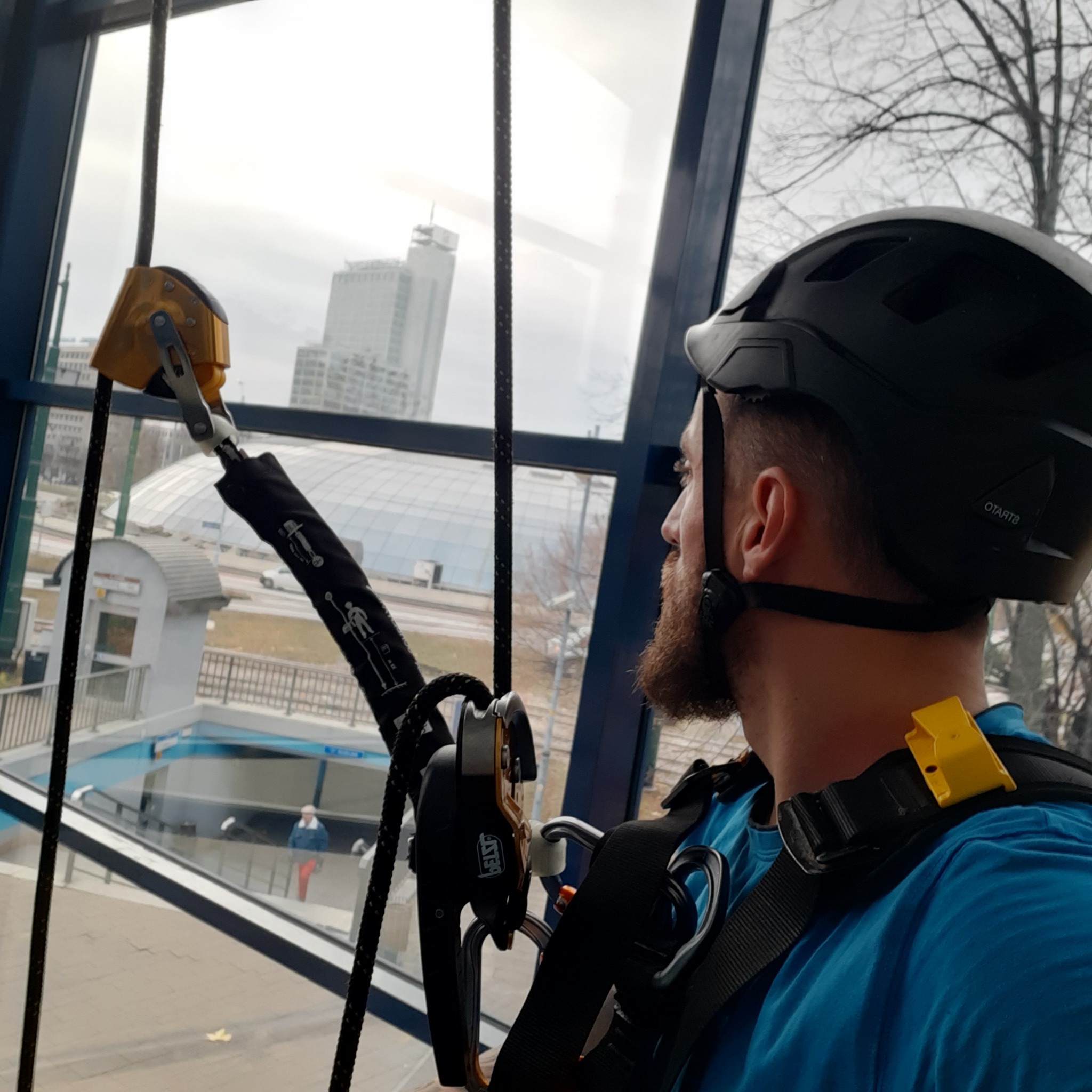 Pracownik w kasku i uprzęży, z narzędziami alpinistycznymi, spogląda przez okno na panoramę miasta. Widoczny fragment wejścia do metra.