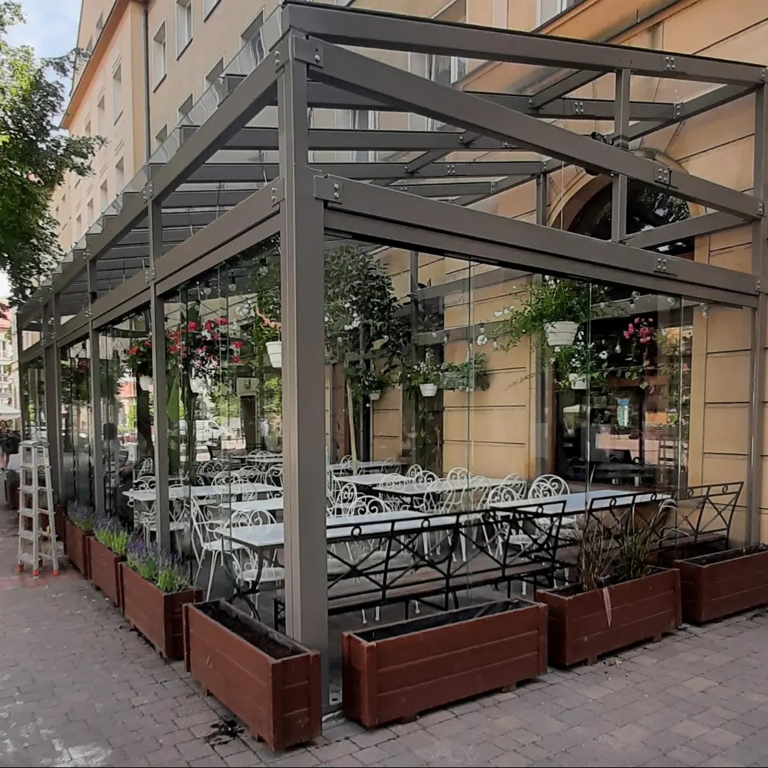 Zadaszony taras restauracji z przeszklonymi ścianami i kwiatami w donicach. Widoczne stoliki i krzesła. Na chodniku drewniane skrzynie z roślinami i drabina.