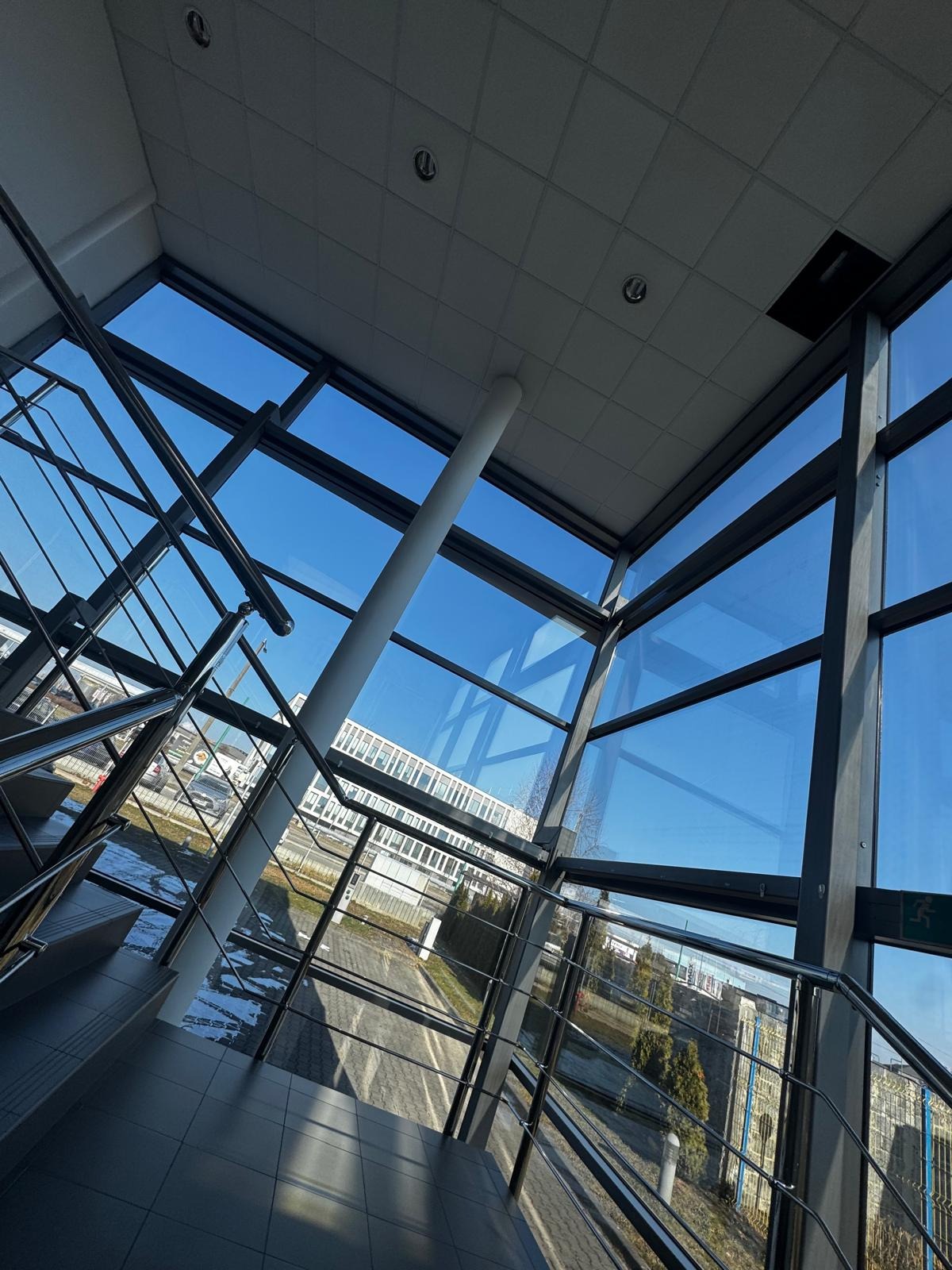 Wnętrze budynku z dużymi oknami, przez które widać błękitne niebo. Metalowe schody i poręcze. Widok na zewnątrz z budynkiem i zielenią. Perspektywa z dołu.