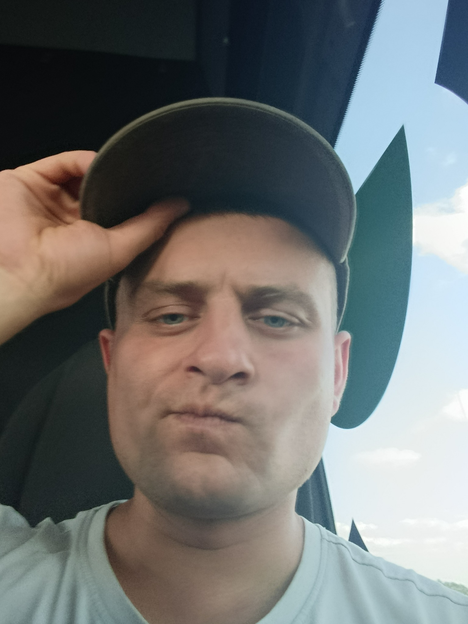 Portret mężczyzny w czapce z daszkiem, z podniesioną ręką do głowy. Twarz o wyrazistych rysach, błękitne oczy. W tle widoczne niebo i fragment wnętrza pojazdu.