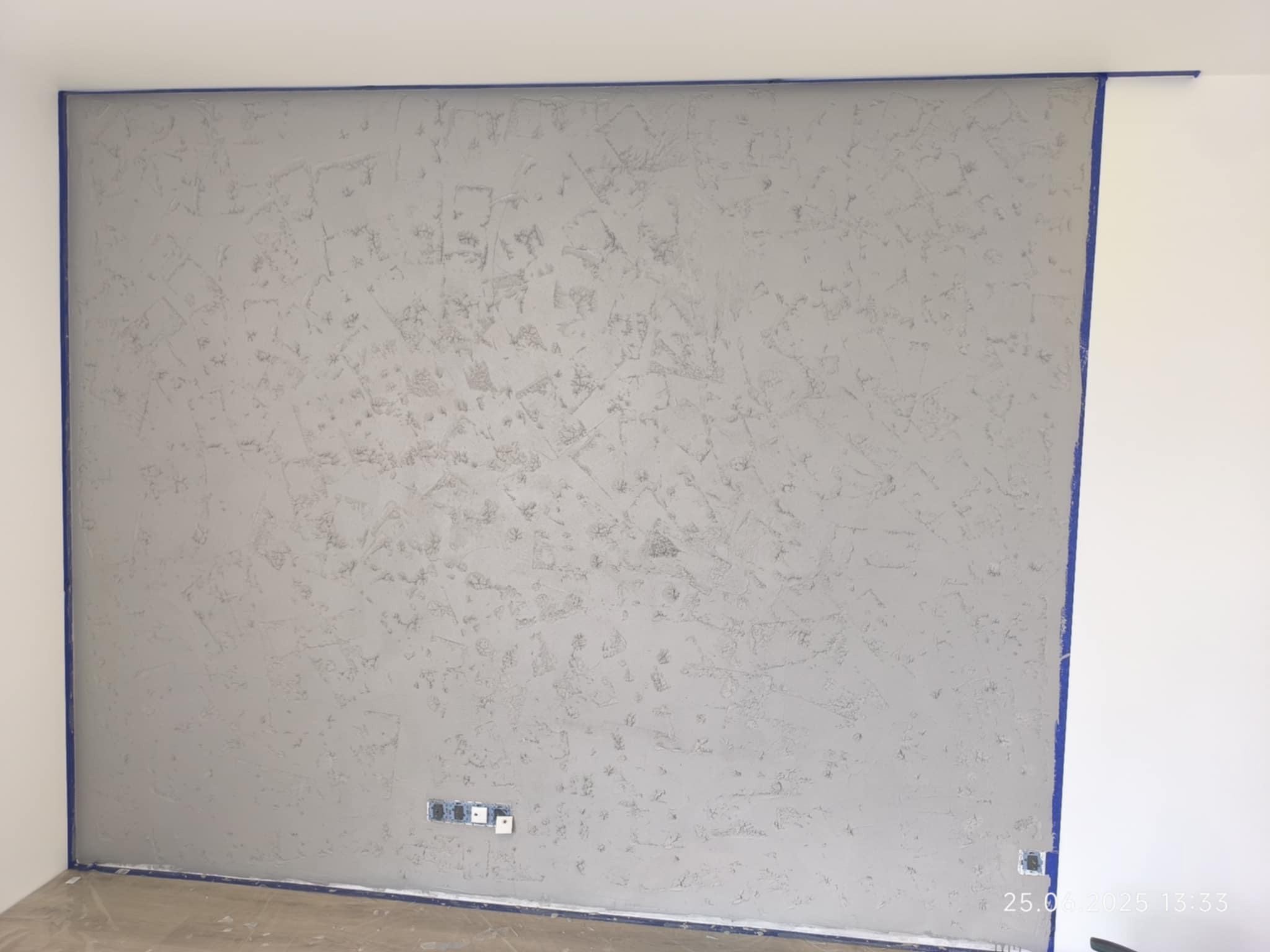 Ściana z efektem dekoracyjnym, zabezpieczona taśmą malarską. Widoczne puszki elektryczne. Szara, teksturowana powierzchnia. Świeżo położona gładź dekoracyjna.