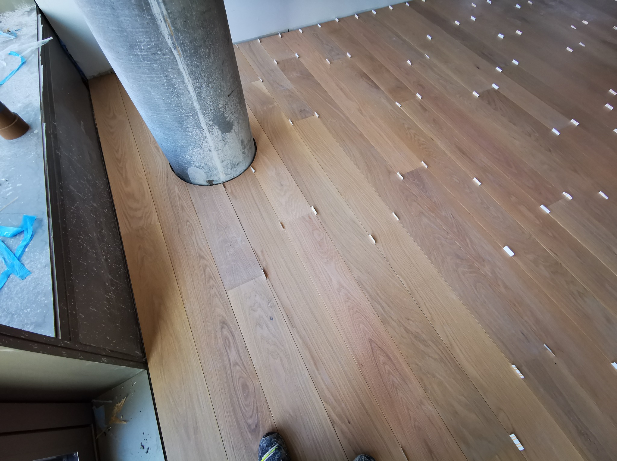 Układanie parkietu w pokoju z kolumną. Widoczne białe kliny dystansowe. Częściowo widoczne buty osoby wykonującej prace. Jasne drewno.