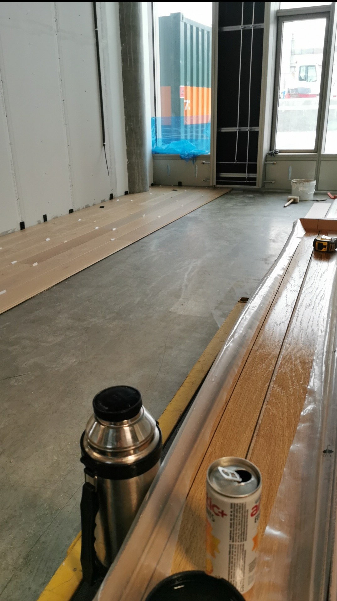 Układanie parkietu w jasnym wnętrzu. Częściowo położone deski, widoczne narzędzia i napój. Duże okna z widokiem na kontener. Etap prac wykończeniowych.