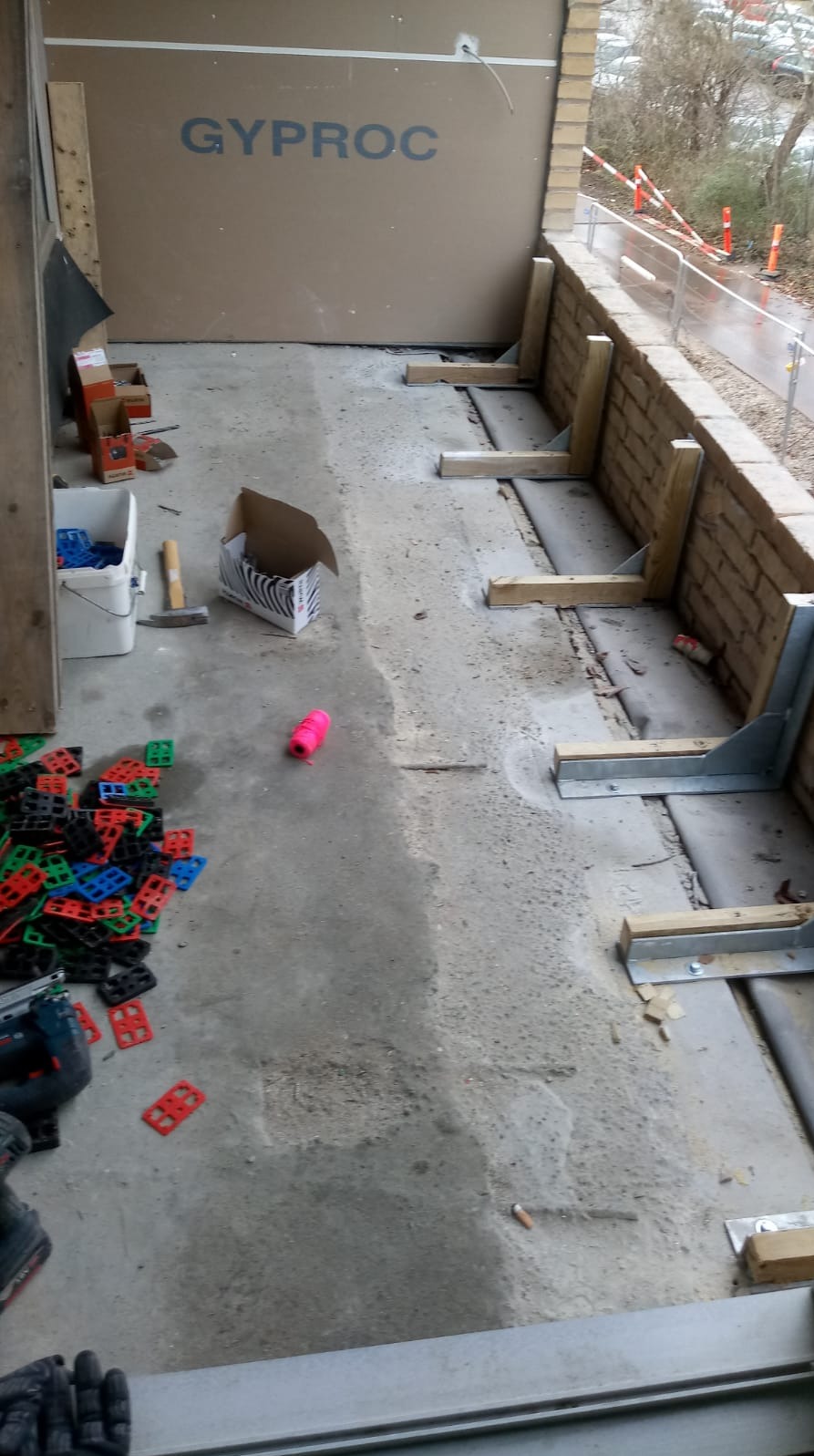 Balkon w trakcie remontu: płyta Gyproc, wsporniki, poziomica, rozsypane krzyżyki glazurnicze, narzędzia. Widok na zewnątrz przez niezabezpieczoną barierkę.