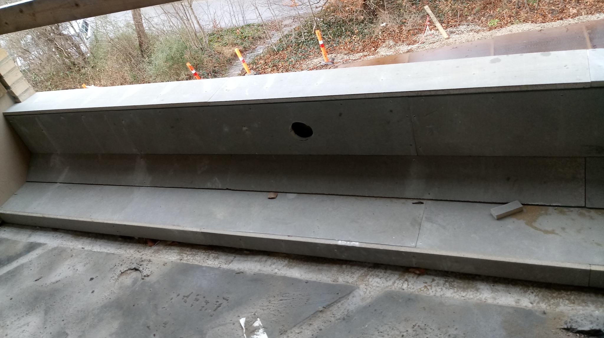 Szara, wykończona wnęka balkonowa z otworem wentylacyjnym, widok z góry. Fragment kostki brukowej leży na dolnej krawędzi. Na zewnątrz widoczne drzewa.