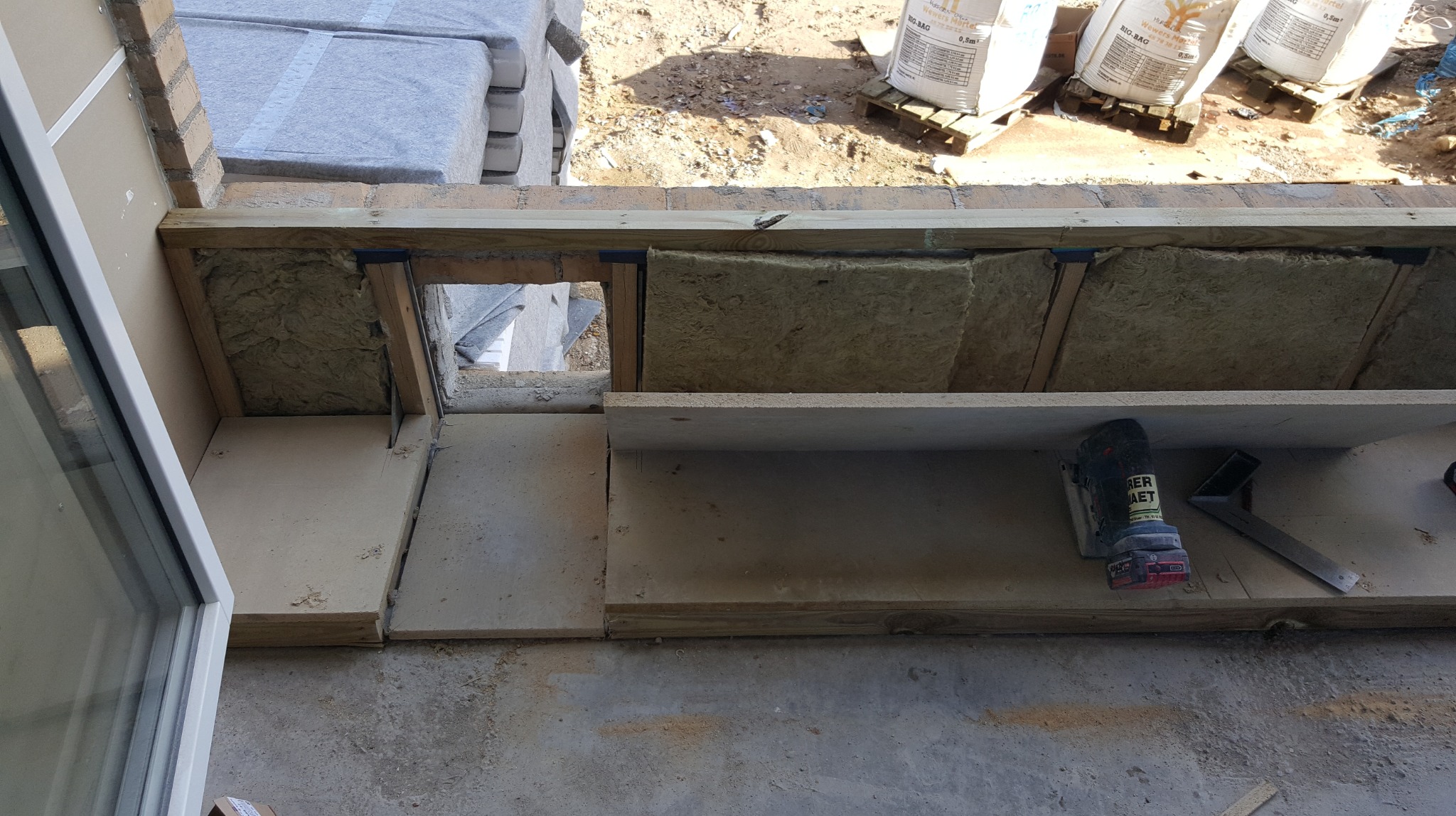 Ocieplenie balkonu wełną mineralną w drewnianej konstrukcji. Widoczne narzędzia i materiały budowlane. Prace wykończeniowe w toku, z zewnątrz widoczne worki z zaprawą.