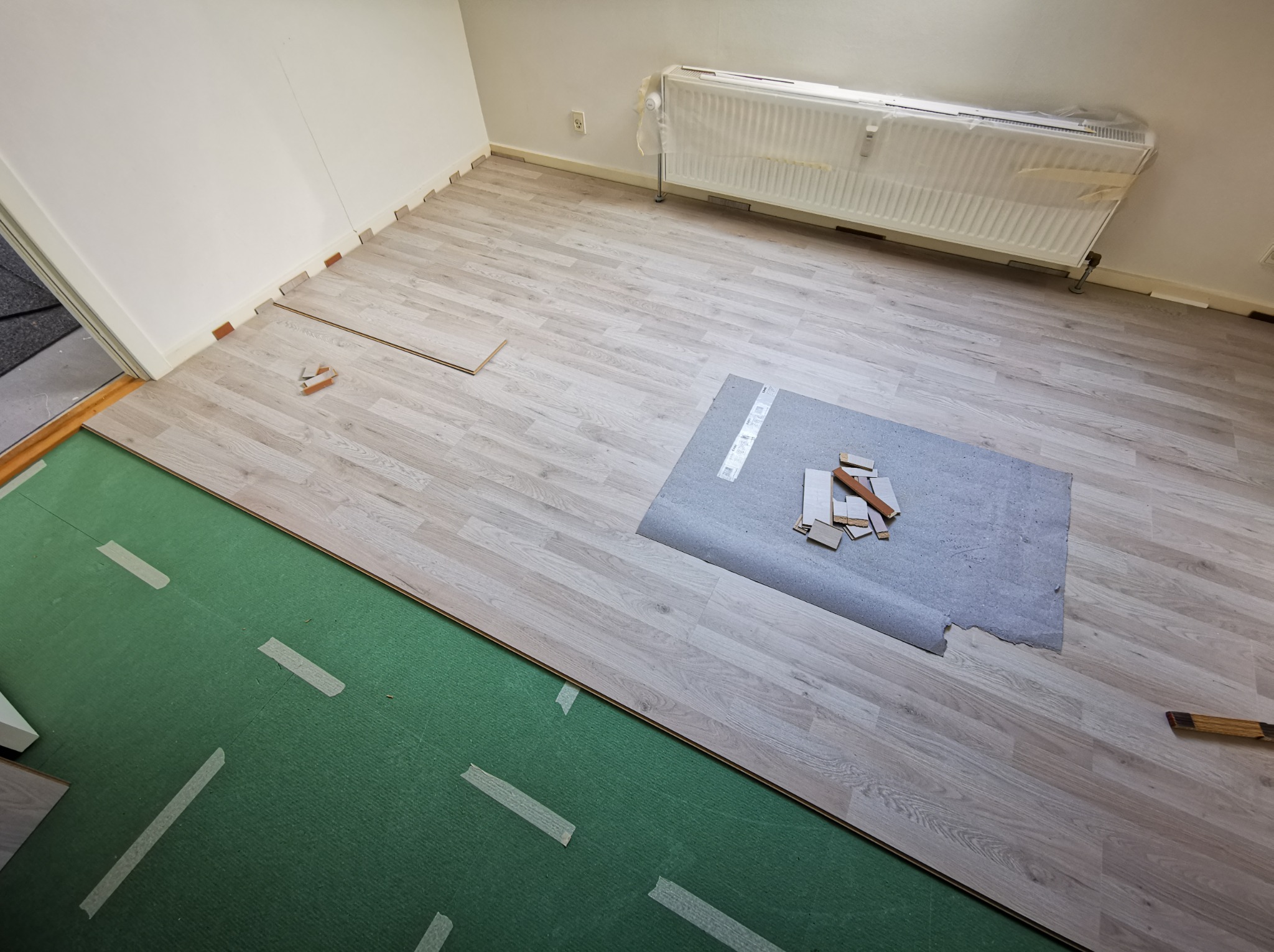 Układanie paneli podłogowych w jasnym odcieniu, widoczna warstwa izolacyjna i akcesoria montażowe. Grzejnik osłonięty folią ochronną.