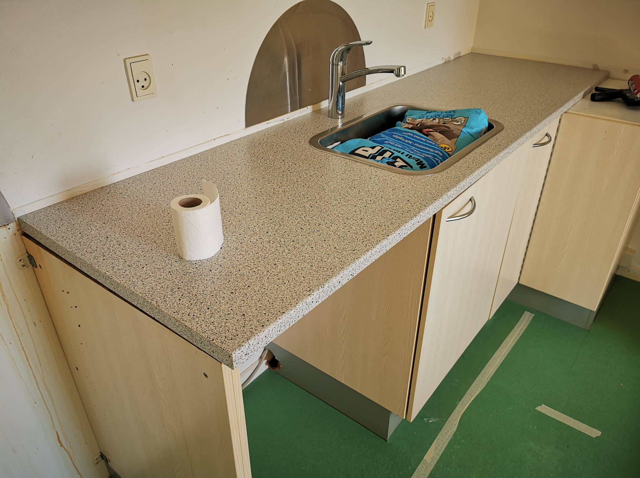 Blat kuchenny z umywalką w trakcie montażu, widoczny worek z zaprawą. Na blacie leży rolka papieru toaletowego, ściany przygotowane do dalszych prac wykończeniowych.