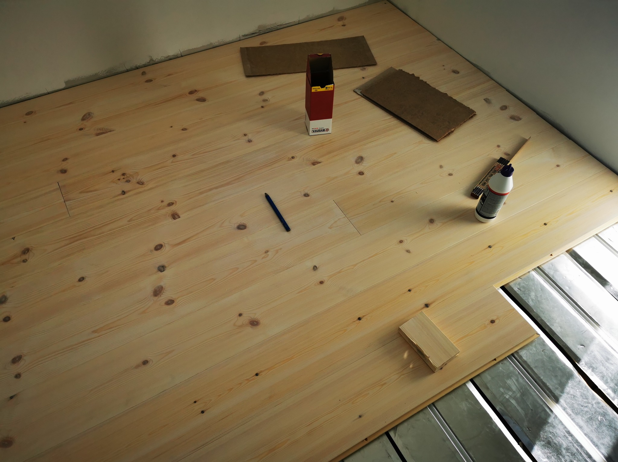 Montaż jasnej, drewnianej podłogi na legarach. Widoczny klej, ołówek i kartonowe podkładki. Część podłogi jeszcze nieułożona, ukazująca metalową konstrukcję.
