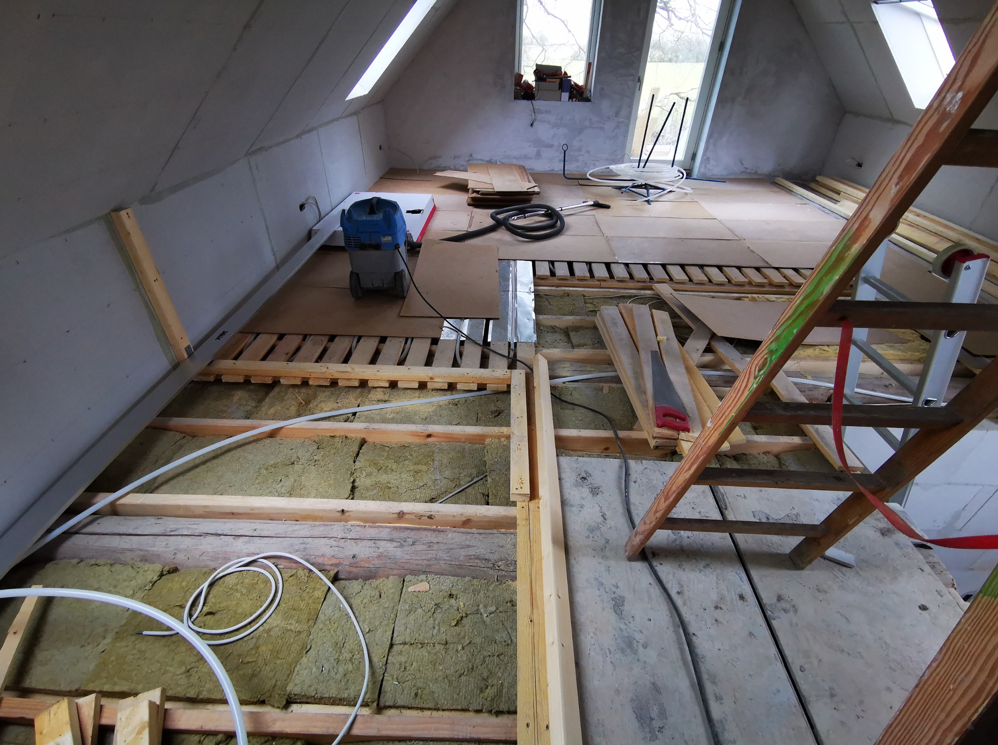 Poddasze w trakcie prac wykończeniowych: ocieplenie wełną mineralną, przygotowanie pod podłogę z płyt. Widoczny odkurzacz przemysłowy, drabina i narzędzia.
