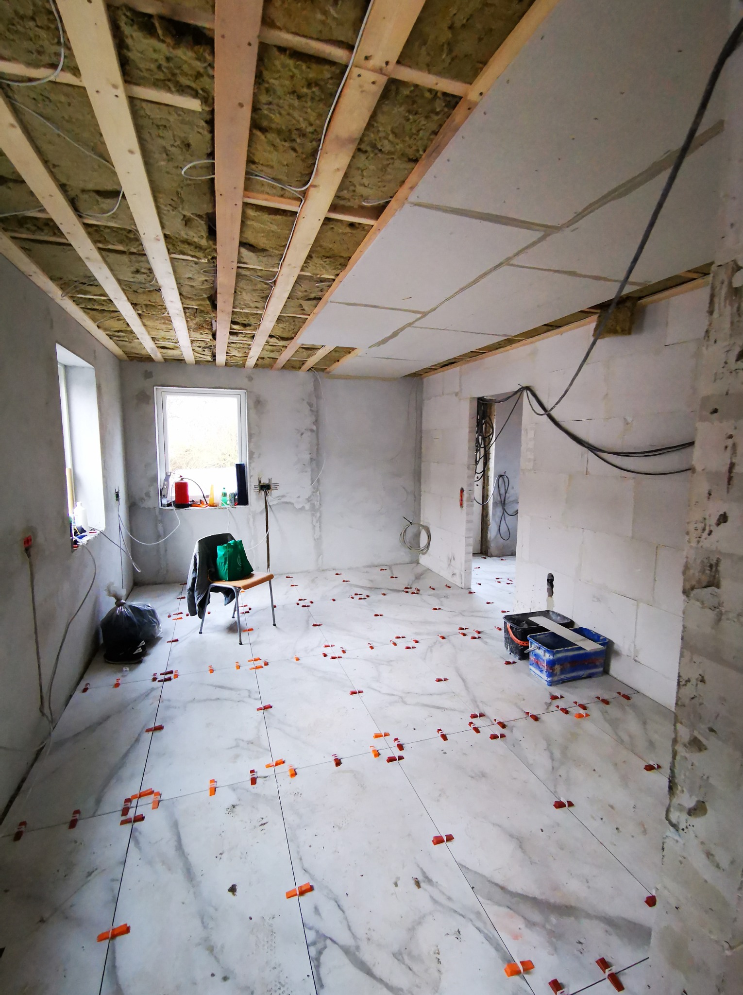 Wnętrze w trakcie prac wykończeniowych: ułożona podłoga z płytek, częściowo wykończony sufit z izolacją, widoczne materiały budowlane i narzędzia.