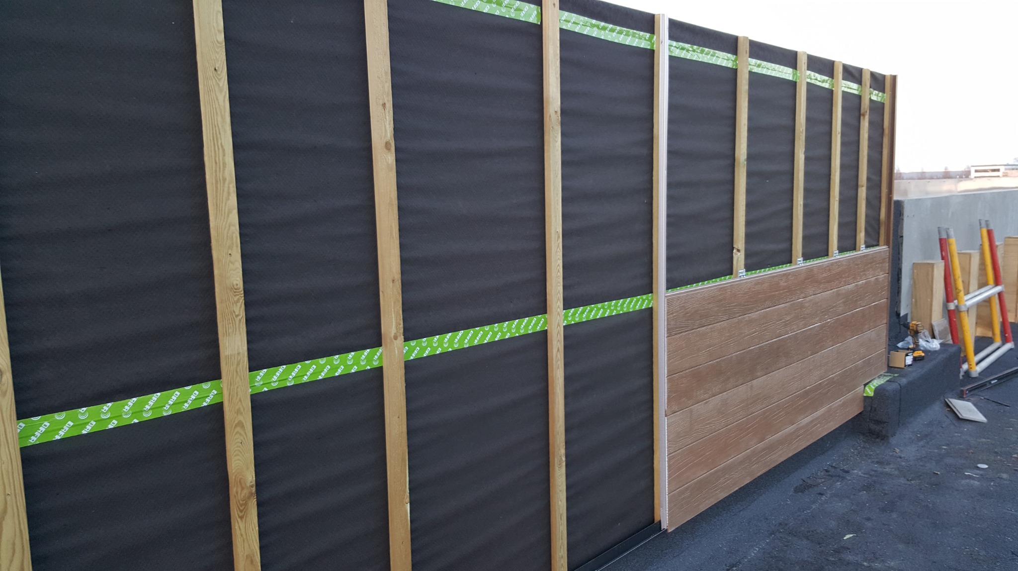 Montaż paneli elewacyjnych na balkonie: widoczne drewniane listwy, czarna membrana i częściowo zamontowane panele w kolorze drewna. W tle drabina i materiały.