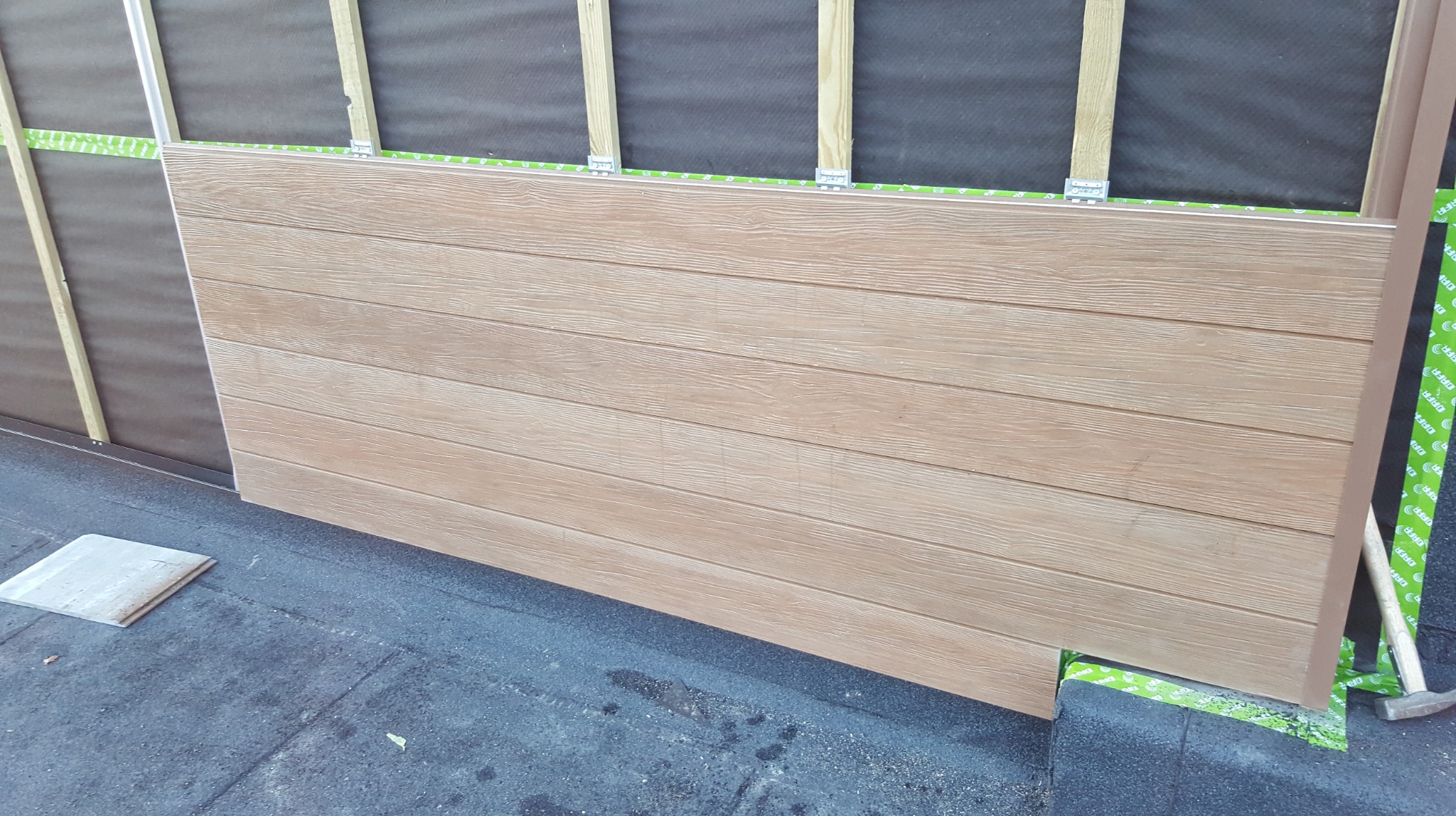 Montaż paneli imitujących drewno na elewacji. Widoczne elementy mocujące, drewniane listwy i fragment dachu pokrytego papą. Prace wykończeniowe w toku.