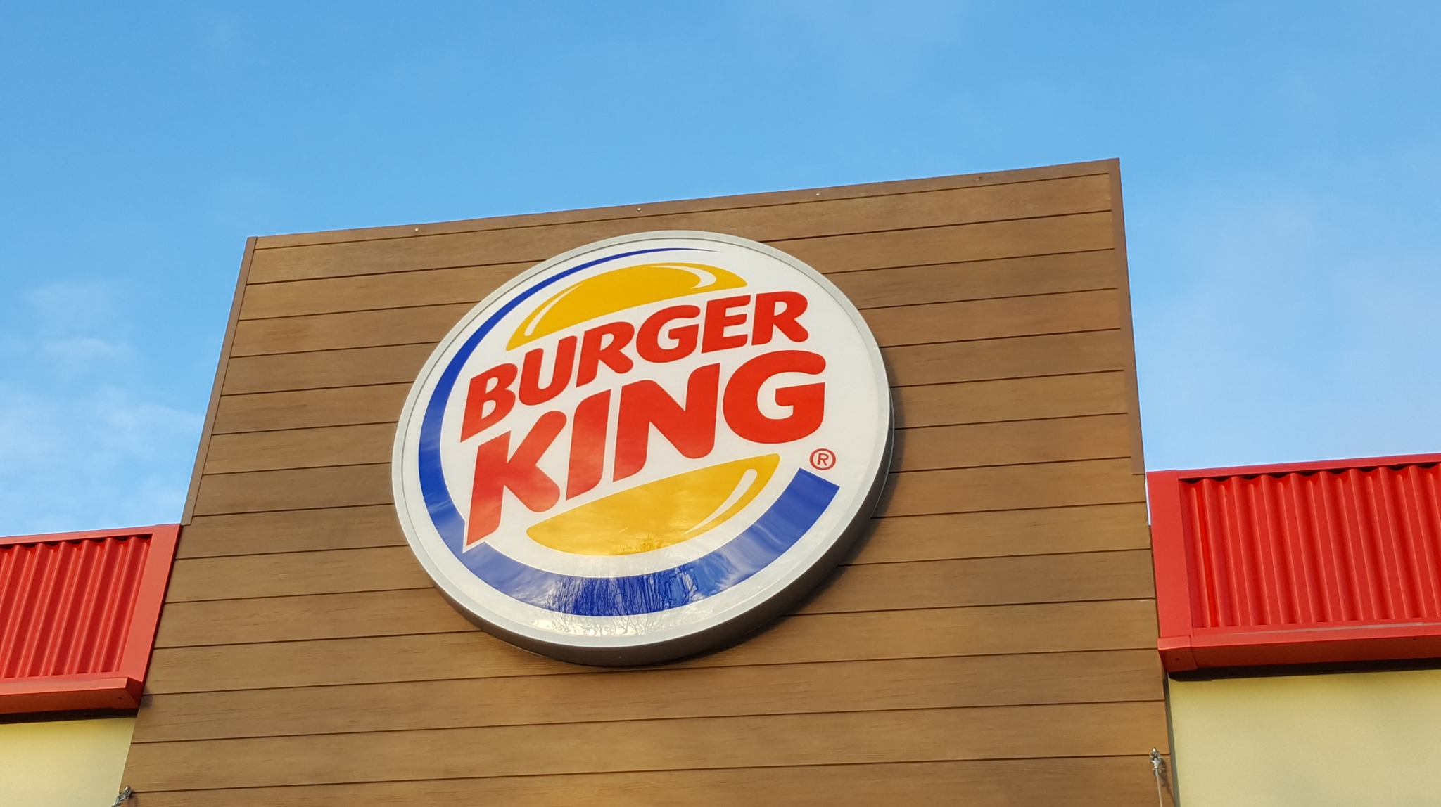 Okrągły szyld Burger King na brązowej elewacji z drewnopodobnymi panelami, widoczny fragment czerwonego dachu i błękitne niebo w tle.