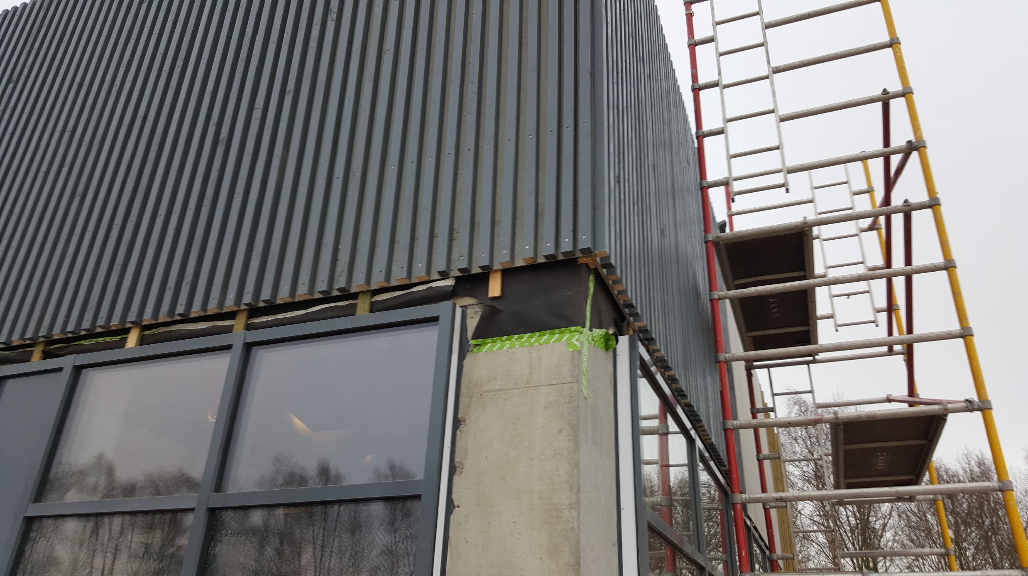 Montaż elewacji z blachy trapezowej na budynku z oknami. Widoczne rusztowanie i zabezpieczenia. Prace w toku, detale mocowania i izolacji.