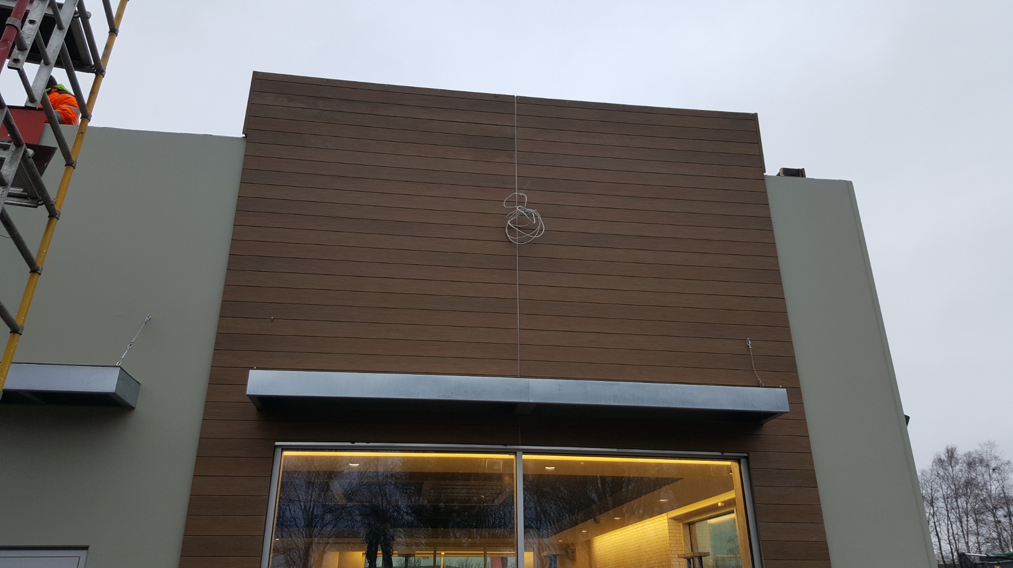 Elewacja budynku z brązową okładziną imitującą drewno, widoczna z dołu. Na ścianie metalowe elementy. Po lewej stronie drabina z pracownikiem.