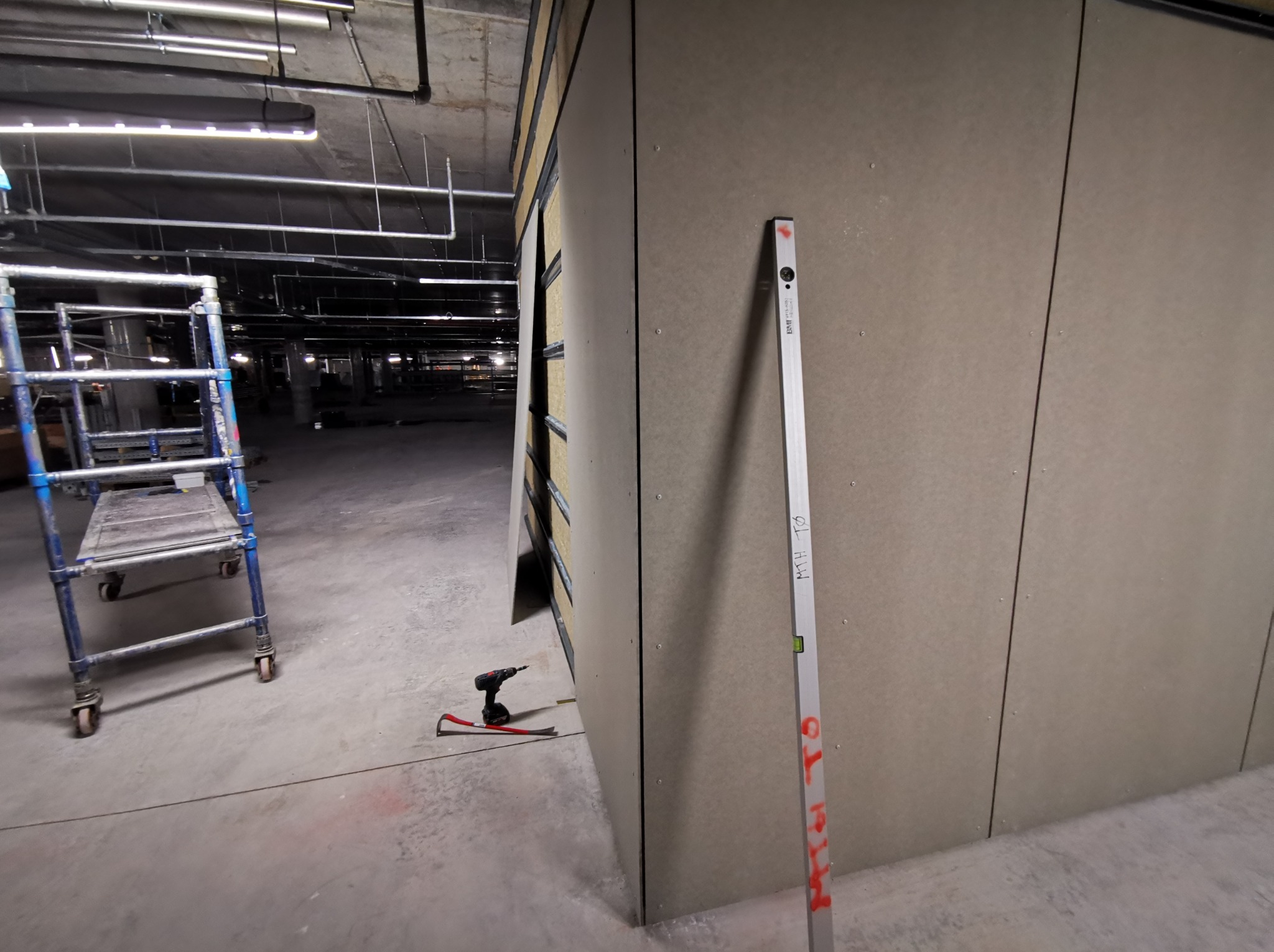 Montaż płyt gipsowo-kartonowych w obiekcie komercyjnym. Widoczny poziomica, wkrętarka i rusztowanie. Surowy stan wykończenia, instalacje pod sufitem.