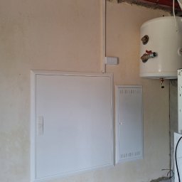 Adam Remontuje - Białe klapy rewizyjne w ścianie, widoczny bojler i rury. Ściana przygotowana do malowania. Szare płytki u dołu ściany. Widoczne niedoskonałości tynku.