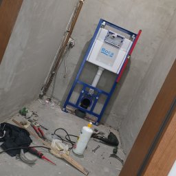 Adam Remontuje - Montaż stelaża podtynkowego WC Roca w surowej łazience. Widoczne narzędzia i materiały budowlane na podłodze, miedziane rury na ścianie.