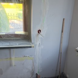 Adam Remontuje - Ściana w trakcie szpachlowania z widocznymi przewodami elektrycznymi i zabezpieczonym oknem folią. Na podłodze folia ochronna, widoczna łata i poziomica.